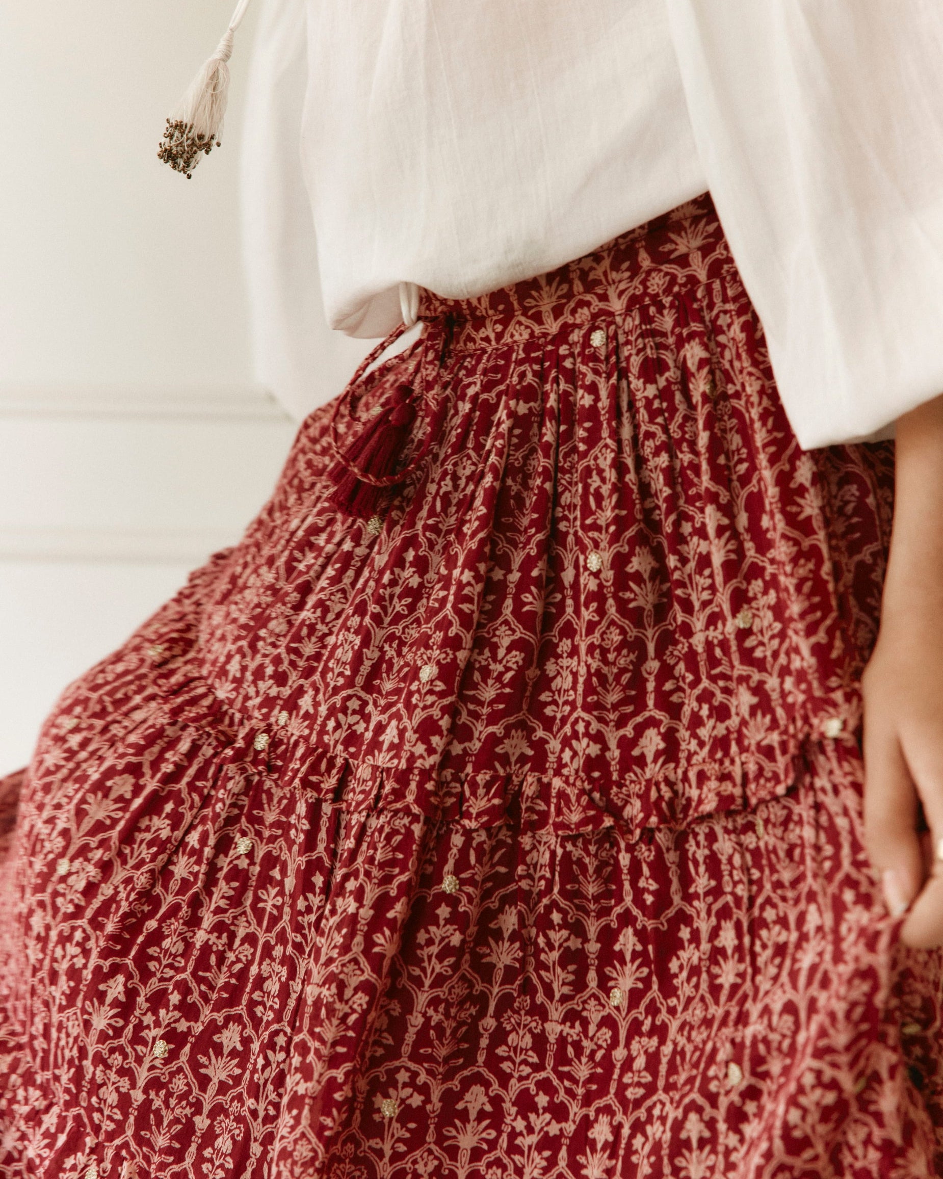 LAKOÈN closeup of a model in our tiered Emile Maxi Skirt, revealing a softly elasticated waistband and tasseled ties. Crafted from fine cotton with a delicate block-inspired print in rose and rich burgundy hues, adorned with shimmering gold embroidered dots. Pictured with our White Emory blouse with hand beaded tassels - slow-luxury - hand-crafted - bohemian - ethically-made