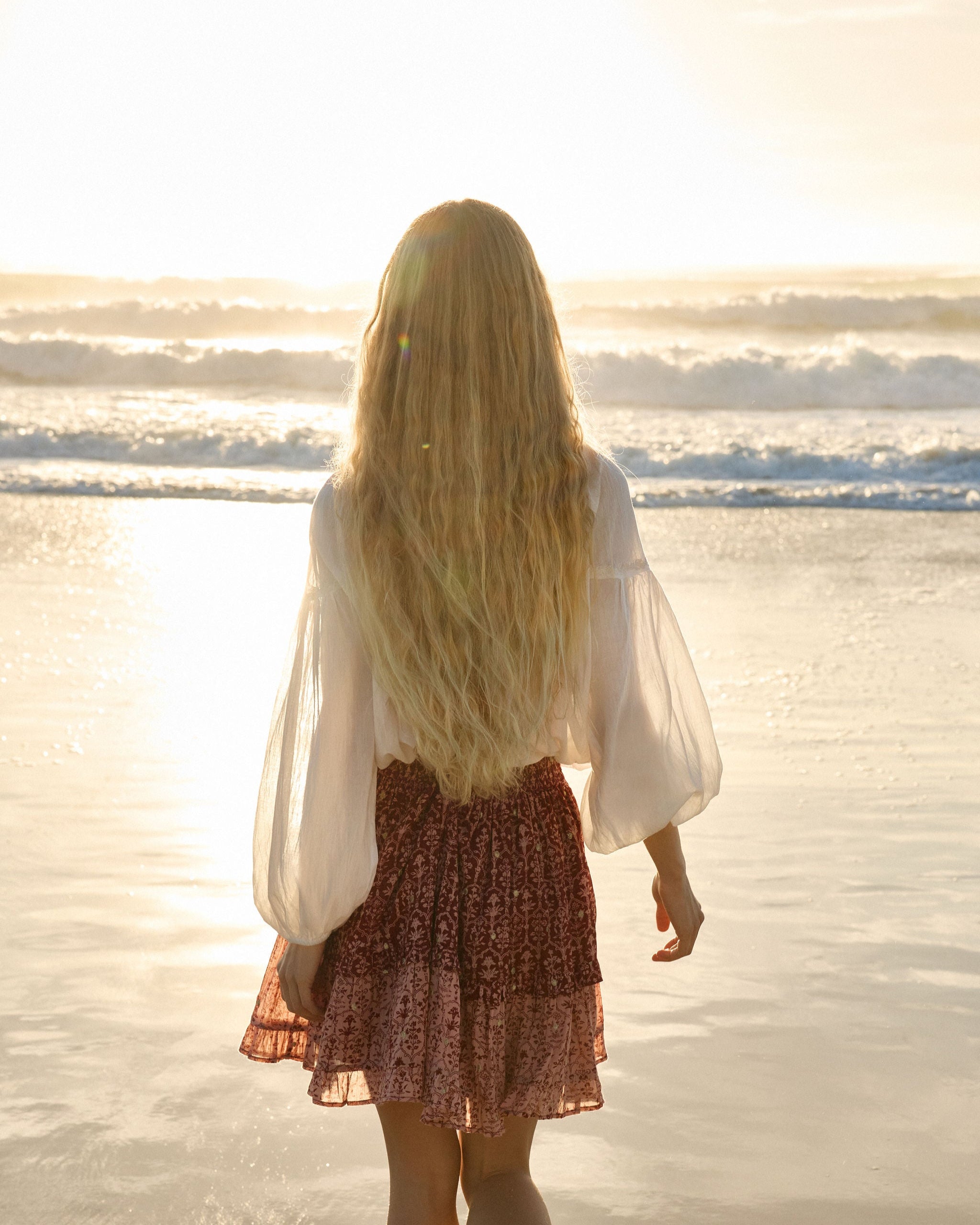 Lakoen model walking towards the ocean in Emory Blouse in soft white cotton with drawstring neckline and waist and 3/4 sleeves and our Emile Mini Skirt in printed cotton, in rose pink & merlot hues, with delicate gold lurex embroidered dots - slow fashion - hand-crafted - ethically made