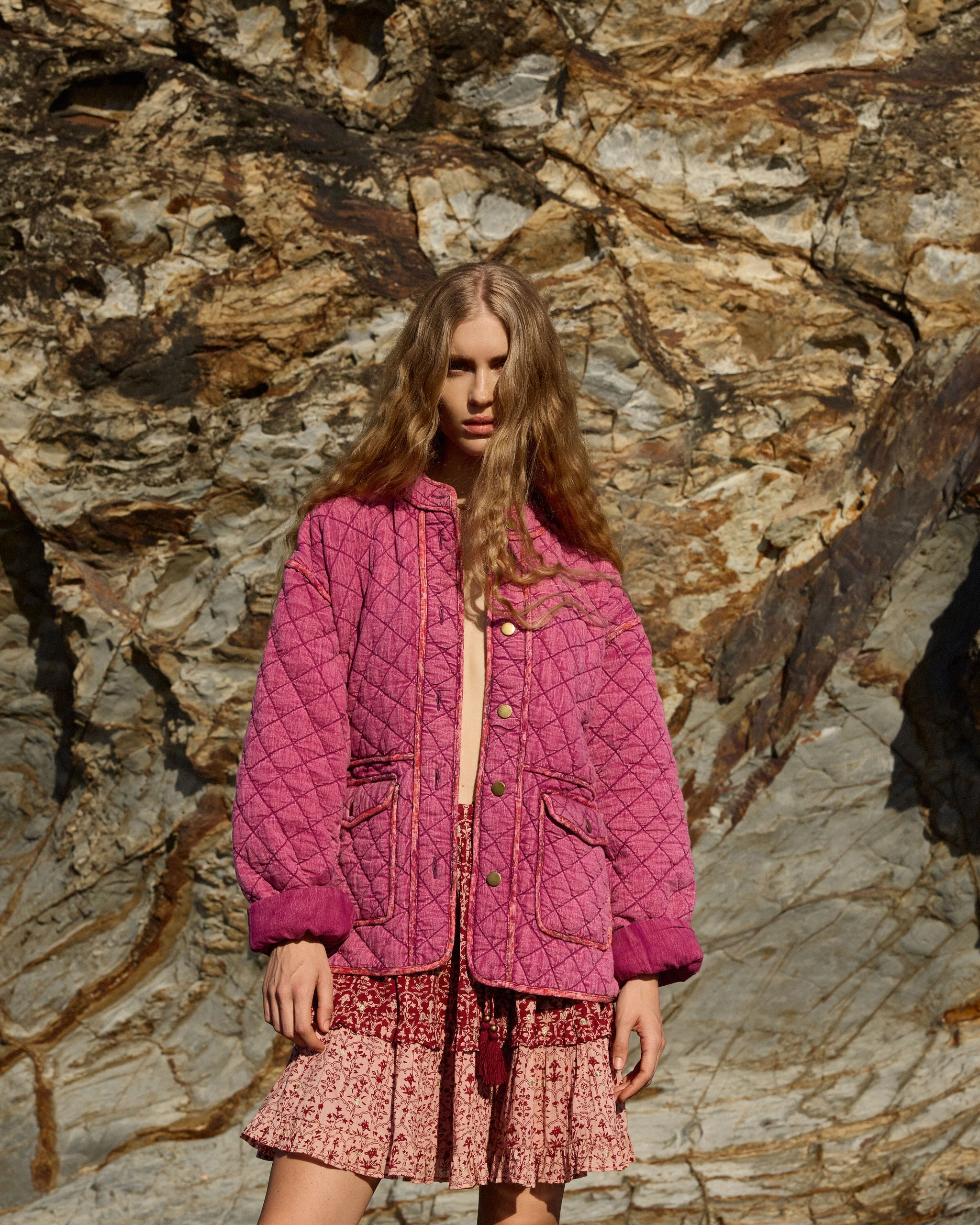 Model in Lakoen clothing posing against sunlit cliff in our Quilted Frankie jacket in washed cerise and our Lakoen Emile Mini Skirt in printed cotton, in rose pink & merlot hues, with delicate gold lurex embroidered dots - slow fashion - hand-crafted - ethically made