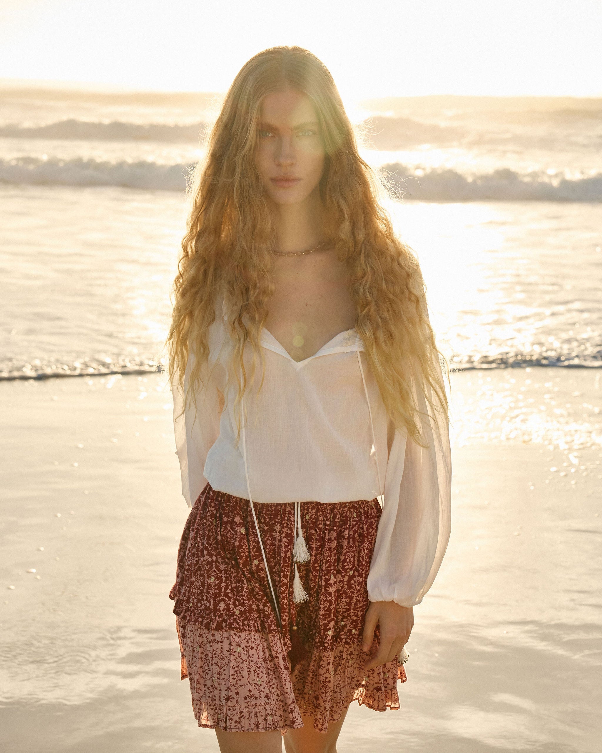 Lakoen model walking away from the ocean in Emory Blouse in soft white cotton with drawstring neckline and waist and 3/4 sleeves and our Emile Mini Skirt in printed cotton, in rose pink & merlot hues, with delicate gold lurex embroidered dots - slow fashion - hand-crafted - ethically made